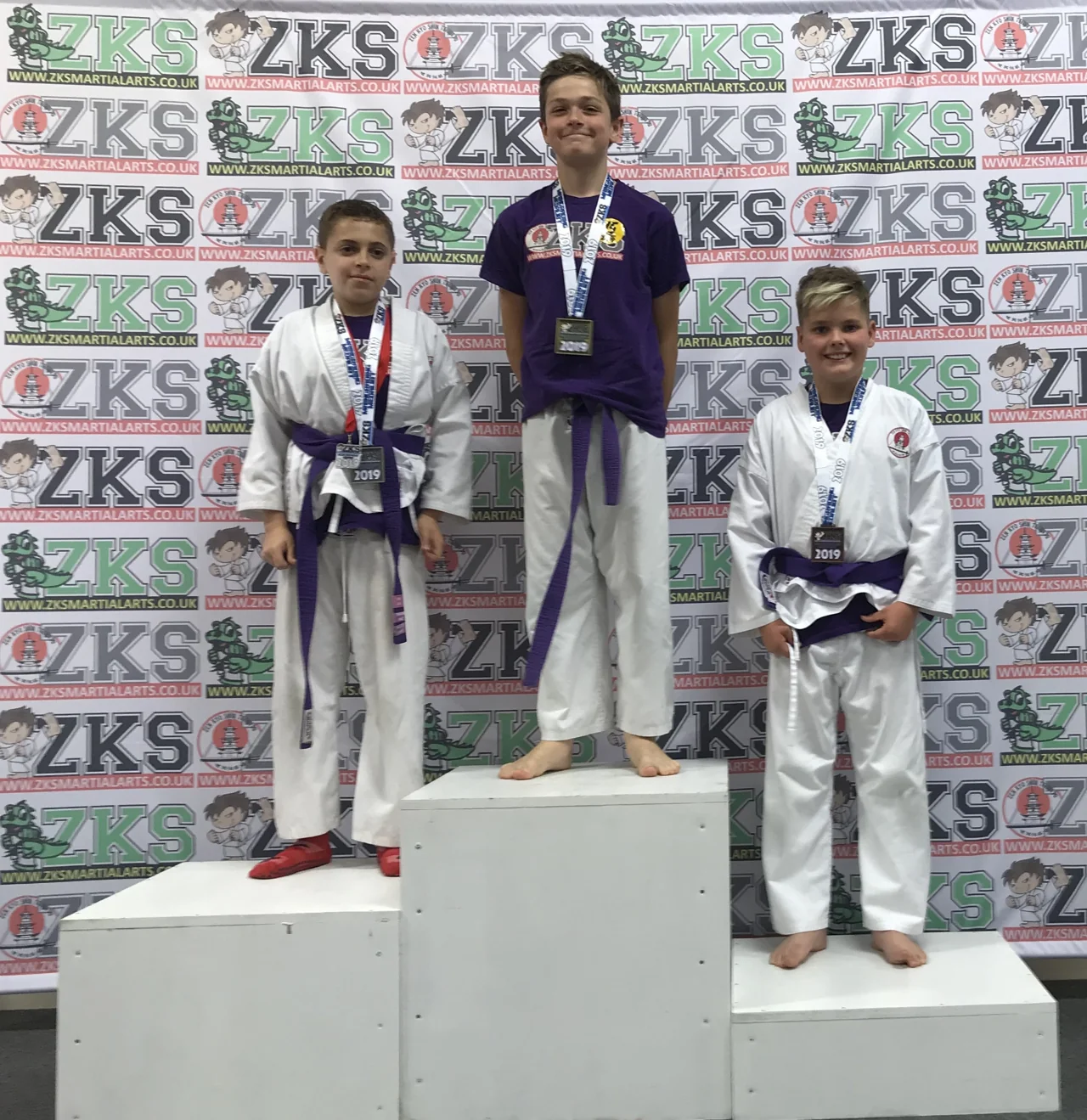 Three Junior MA purple-belt students wearing medals on a ZKS podium